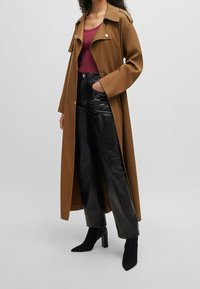 Dlouhý hnědý trench coat s velkými klopami, zkombinovaný s vínovým topem, černými lesklými širokými kalhotami a černými vysokými kotníkovými botami.