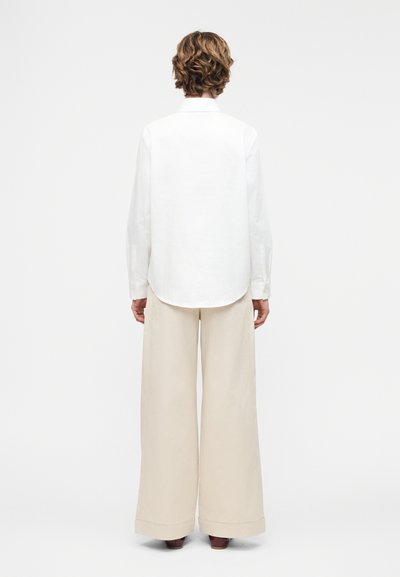 Άτομο που στέκεται με την πλάτη γυρισμένη, φορώντας άσπρο πουκάμισο με μακριά μανίκια και μπεζ παντελόνι με φαρδιά πόδια, μπροστά σε απλό ανοιχτό φόντο.