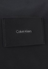 Czarna torba Calvin Klein z teksturowaną powierzchnią materiału i gładką skórzaną naszywką z nazwą marki wytłoczoną na srebrno.