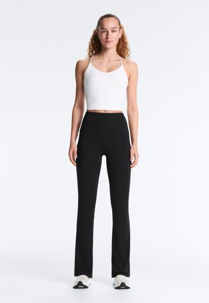 Tânără femeie în picioare, orientată frontal, purtând un top crop alb fără mâneci, colanți negri cu talie înaltă și adidași albi pe un fundal simplu.