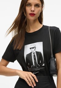 Must tükk mustast puuvillast särk, millel on graafiline print mehega prillide ja ülikonna seljas. Lühikesed varrukad ja ümar kaelus. Sobitub musta kotiga.