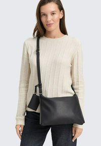 Schwarze Leder-Crossbody-Tasche mit strukturierter Oberfläche, verstellbarem Trageriemen und kompakter Form. Das Modell trägt einen hellen, cremefarbenen, gerippten Pullover.
