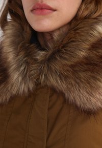 Brun ytterklädsel med en bred, fluffig fuskpälskrage i nyanser av ljus och mörkbrunt. Mjuk tygstruktur under kragen.