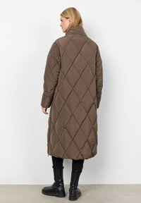 Bruine gequilte lange jas met een hoge kraag, voorzien van een diagonaal ruitpatroon en zijzakken, gecombineerd met zwarte enkelboots.