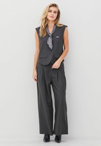 Sacou gri fără mâneci cu un buzunar și pantaloni largi plisati asortați, completat de o cravată gri și cizme negre până la glezne.