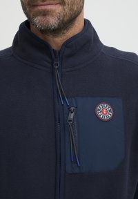 Marinefarget fleecejakke med høy krage, frontlukking med glidelås, blå stofflomme og et rundt logo-patch. Myk tekstur, funksjonelt design.