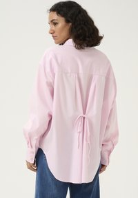 Lyserød oversized skjorte med knapper og spidse krave, lange ærmer og en samlet detalje bagpå, der er bundet om taljen. Denimbundkort synlige.