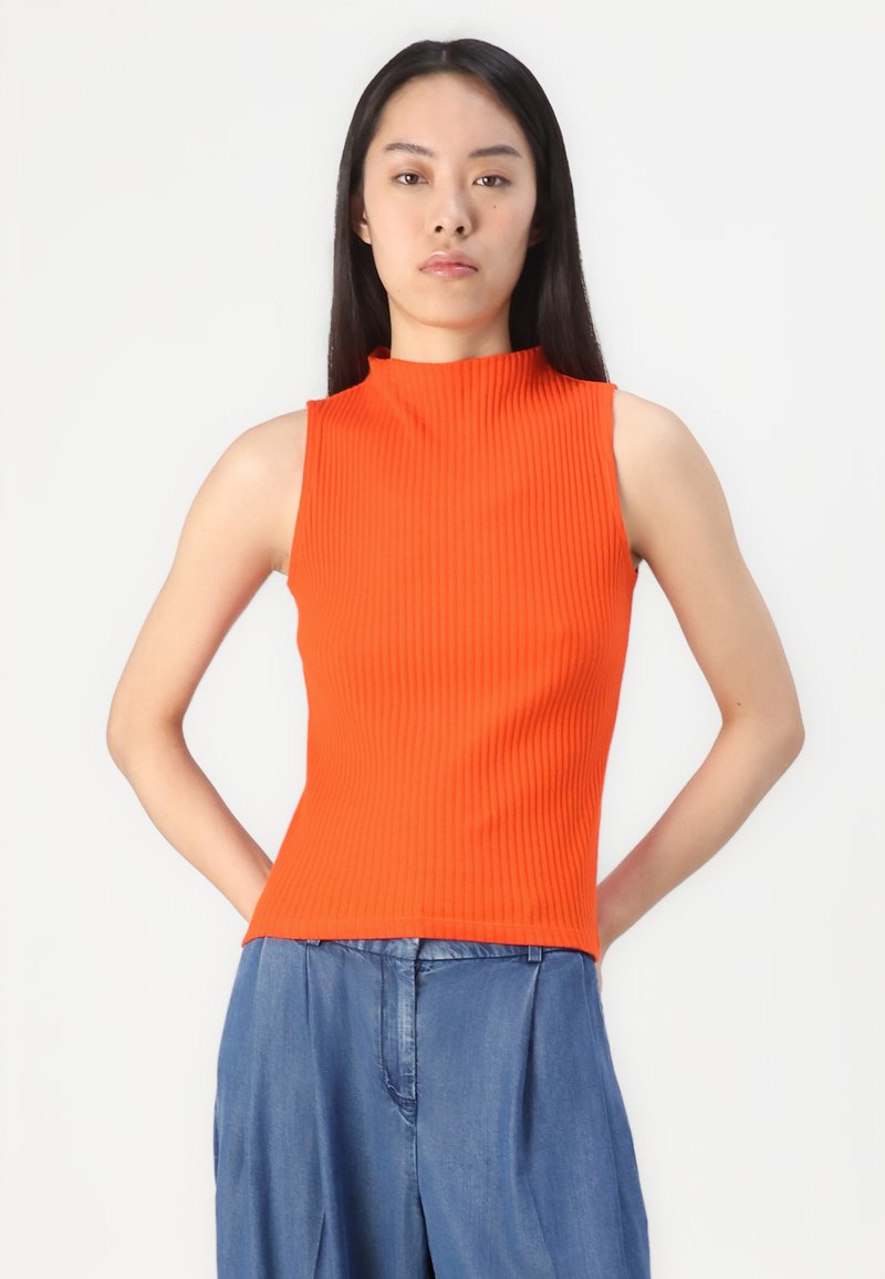 Top orange côtelé sans manches avec un col haut, de forme ajustée et en tissu texturé. Porté avec un pantalon en denim bleu clair ample.