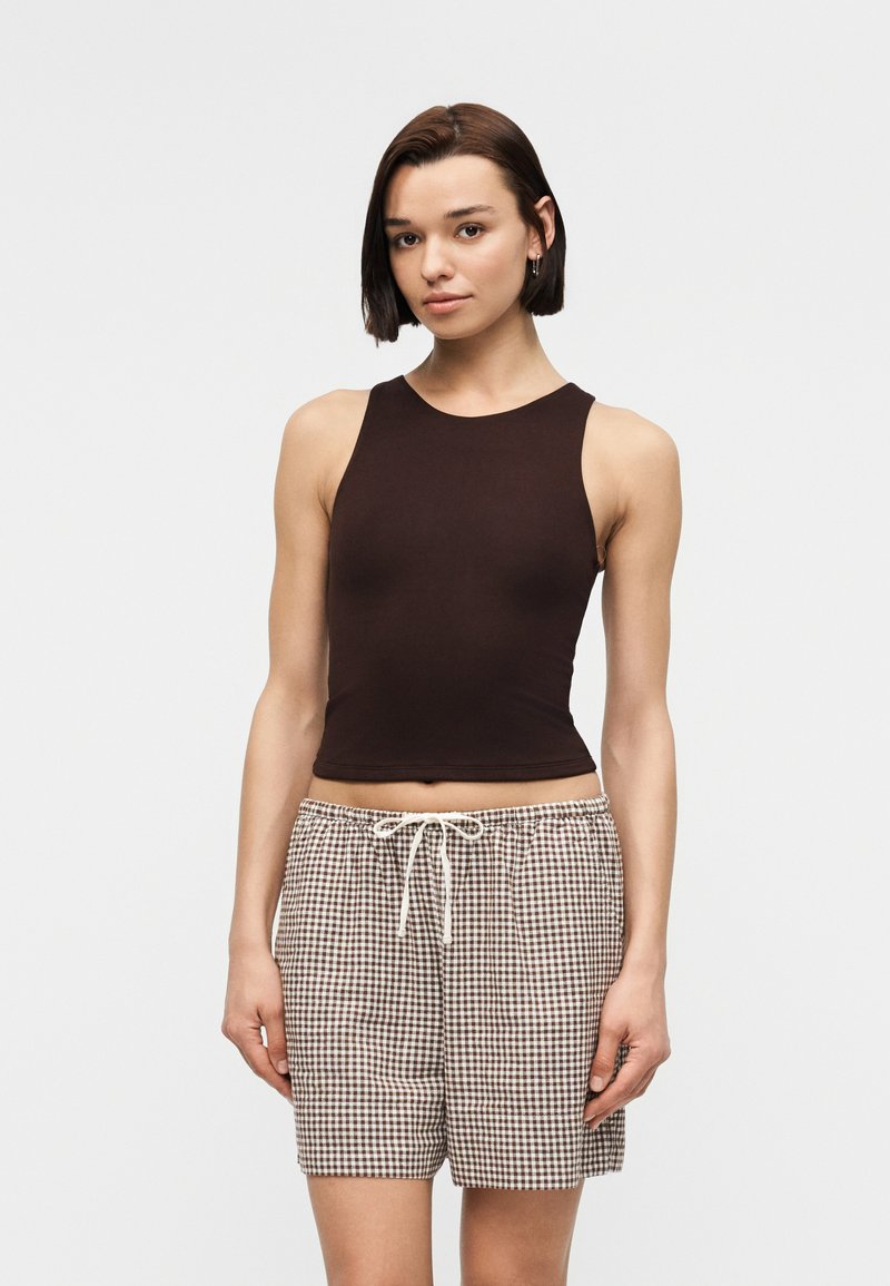 Jeune femme portant un haut sans manches marron foncé et un short à carreaux vichy taille haute avec cordon de serrage, debout devant un fond uni.