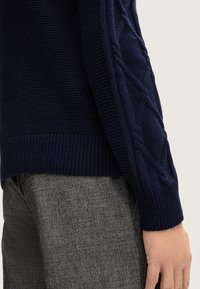 Marineblauer Strickpullover mit Kabelmuster an den Ärmeln, getragen mit grauen Fischgrätenhosen, wobei das Handgelenk und die Hand entspannt an der Seite gezeigt werden.