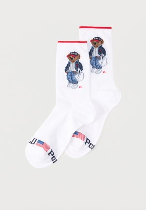 Hvide sportssokker med rød kant, der viser en bjørn iført en Polo-kasket, jakke, jeans, sneakers, der holder en hvid taske, og et amerikansk flag nær tæerne.