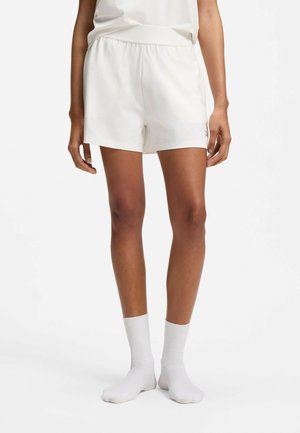 Femme portant un short blanc à taille élastique, chaussettes blanches, debout avec les bras détendus le long du corps sur un fond uni.