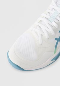 Scarpa sportiva bianca con suola e dettagli di colore azzurro chiaro, tomaia in rete e lacci bianchi, mostrata dall'alto, con un focus sulla punta e sull'area dei lacci.