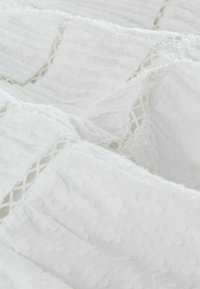 Tissu blanc avec des rayures texturées et des points en relief. Les bords présentent des accents de dentelle détaillés, ajoutant une dimension au matériau.