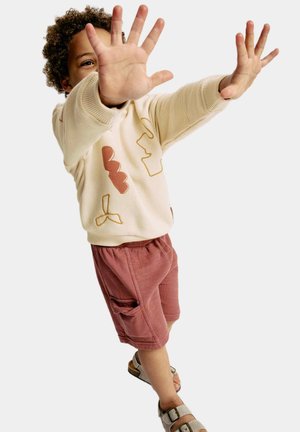 Maglione beige con motivi astratti in ruggine e giallo, abbinato a pantaloni pantaloni rosa con una tasca laterale. Sandali marrone chiaro.