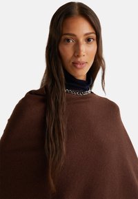 Femme aux longs cheveux bruns portant un poncho en tricot marron par-dessus un col roulé noir, accessoirisé d'un collier en chaîne argentée, sur un fond blanc.