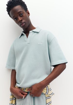 Fiatal férfi rövid, göndör hajjal, világoskék pólóingben, csíkos sárga-kék ruhadarabot igazít deréknál, fehér háttér előtt.