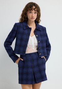 Marinblå ruta tweedjacka med metalliska detaljer och strukturerad krage, i kombination med en matchande kjol. Spetstopp under jackan ger textur.