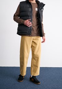 Schwarze Steppweste über einem braunen Langarmshirt, kombiniert mit beigen weiten Hosen und schwarzen Sneakers mit braunen Akzenten.