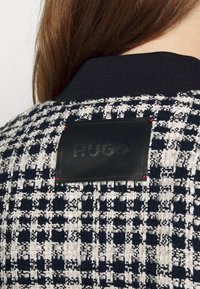 Personne aux cheveux bruns portant une veste à motif pied-de-poule noir et blanc avec une étiquette noire "HUGO" cousue dans le haut du dos.