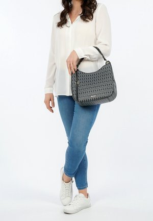 Frau in weißer Bluse und blauen Jeans, die eine graue gemusterte Schultertasche mit schwarzem Trageriemen hält, und weiße Sneakers trägt.