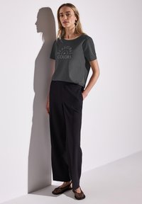 Graues Kurzarm-T-Shirt mit "SEASONAL COLORS" in Weiß bedruckt, kombiniert mit schwarzen weiten Hosen und schwarzen Mesh-Slippern.