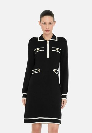 Robe en tricot noire à manches longues, avec des accents blancs, un col zippé, et deux poches avant ornées de boutons dorés et de détails de logo.