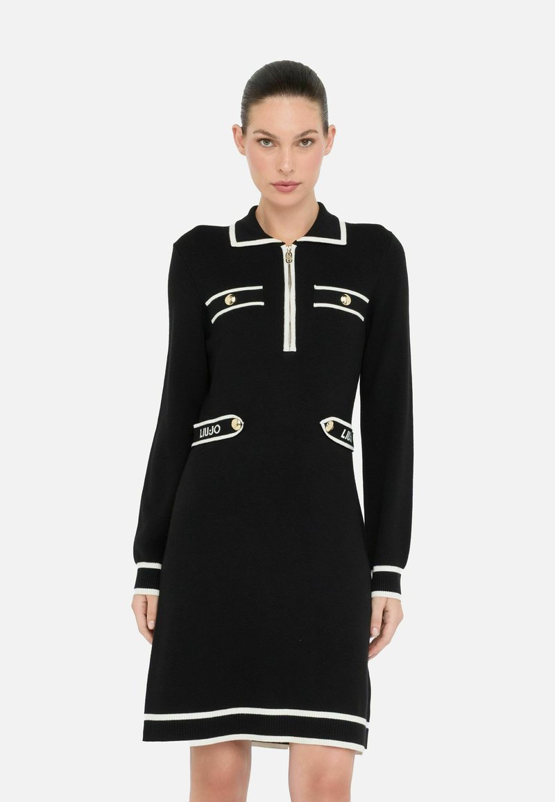 Robe en tricot noire à manches longues, avec des accents blancs, un col zippé, et deux poches avant ornées de boutons dorés et de détails de logo.