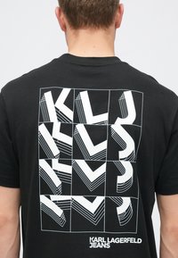 Juodas medvilninis marškinėlis, kuriame matoma drąsi, geometrinė balta grafika su pakaitinėmis raidėmis „K“, „L“ ir „J“ bei sluoksniuotomis vizualinėmis struktūromis.