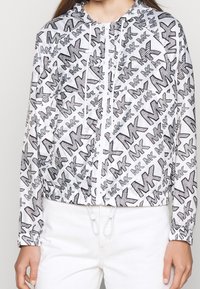 Biała kurtka z powtarzającym się czarnym wzorem stylizowanych liter i logo, z zamkiem błyskawicznym z przodu, ściągaczem w talii oraz kapturem.