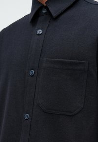 Dunkelblaue Hemdbluse aus einem weichen, strukturierten Stoff, mit einem spitzen Kragen und einer einzelnen Brusttasche mit abgerundetem oberen Rand.