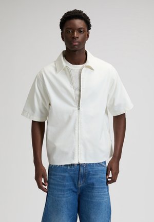 Άνδρας με ανοιχτόχρωμο κοντομάνικο πουκάμισο με φερμουάρ πάνω από πλεκτό πουλόβερ και φαρδιά μπλε τζιν, όρθιος μπροστά σε ανοιχτό γκρι φόντο.