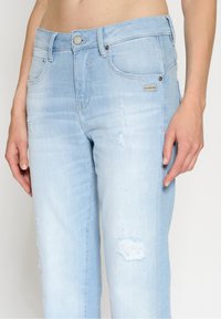 Ljust blå denimjeans med en avslappnad passform, slitna detaljer och en knappstängning i midjan. Diskret varumärkesetikett synlig på framsidan.