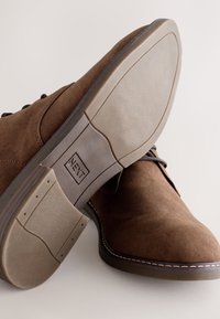 Braune Wildlederschuhe mit abgerundeter Spitze, schwarzen Schnürsenkeln und einer strukturierten grauen Gummisohle. Verfügt über kontrastierende Nähte und ein Logo auf der Sohle.