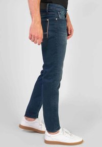 Jeans in denim blu scuro con una vestibilità slim, dotati di tasche davanti e dietro, dettagli di cucitura chiara e una texture liscia. Indossati con sneakers bianche.