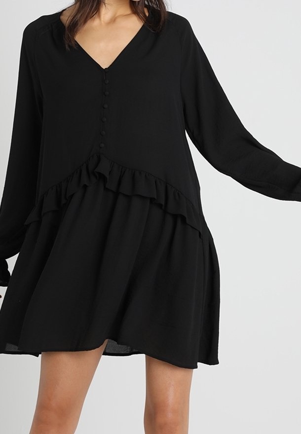 Femme portant une robe noire ample à manches longues, avec des boutons et des détails à volants autour de la taille et de l'ourlet de la jupe.