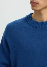 Hombre vistiendo un suéter de cuello redondo acanalado azul, mostrando los hombros, cuello y parte de la parte inferior de la cara con una expresión neutral.