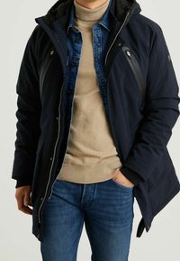 Granatowa kurtka z kapturem, zamkiem z przodu i kieszeniami na piersi. Nałożona na beżowy golf i dżinsową koszulę, zestawiona z niebieskimi dżinsami.