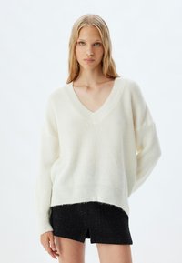 Weißer Strickpullover mit V-Ausschnitt, geripptem Muster und überschnittenen Schultern, kombiniert mit schwarzen strukturierten Shorts.