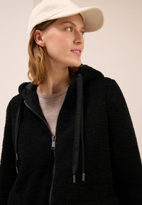 Jeune femme portant une casquette beige, une veste en polaire noire à zip et un haut beige, regardant vers sa droite devant un fond uni.