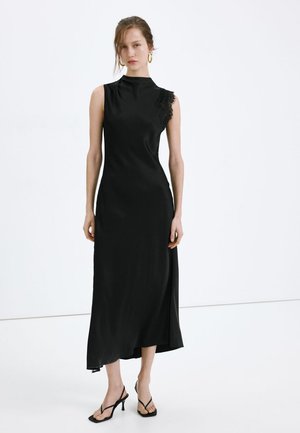 Mujer que lleva un vestido midi negro sin mangas con detalle de encaje en un hombro y sandalias negras de tacón con tiras, de pie sobre un suelo blanco.