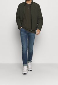 Olivgrön jacka med hög krage över en mörk olivgrön henleytröja, kombinerad med blå jeans och vita sneakers. Avslappnad passform och minimalistisk design.