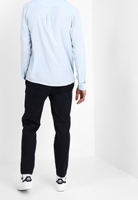 Chemise bleu clair à manches longues avec un pli au dos, associée à un pantalon sombre et des baskets blanches avec des accents noirs.