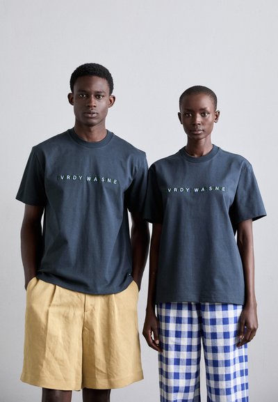 Dunkelgraue Baumwoll-T-Shirts mit gesticktem grünem Text, kombiniert mit beigen Shorts und blau-weißen karierten Hosen, an zwei stehenden Modellen.