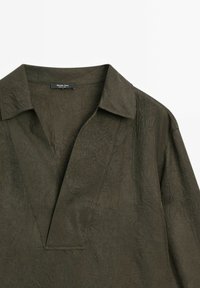 Oliwkowa koszula z kołnierzykiem o pomarszczonej fakturze; ma dekolt z listwą i subtelne detale przeszycia. Metka czyta "Massimo Dutti."