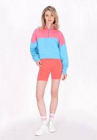 Sudadera con cremallera de dos tonos colorida en azul claro y rosa combinada con pantalones cortos de ciclismo rosas. Zapatillas blancas y calcetines a rayas completan el look.