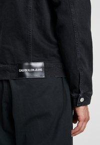Černá džínová bunda s matnou nášivkou s logem "CALVIN KLEIN JEANS", kovovými knoflíky a kontrastním stehováním podél švů.