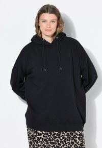Žena s krátkými vlnitými vlasy má na sobě černou oversize mikinu a sukni s leopardím potiskem, stojí na bílém pozadí s rukama za zády.