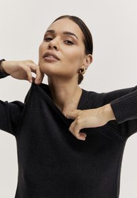 Črni pleteni pulover z širšim izrezom in rebrastimi manšetami. Model nežno vleče za ovratnik, kar prikazuje mehko teksturo blaga.