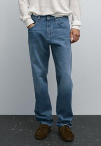 Jeans in denim azzurro chiaro con vestibilità dritta, cinque tasche e dettagli cuciti, indossati con scarpe in suede marrone e un maglione color crema.